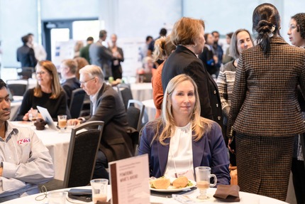 A woman seated at a table looks at the camera and smiles. Behind her is a room filled with more tables and groups of people engaged in conversations.