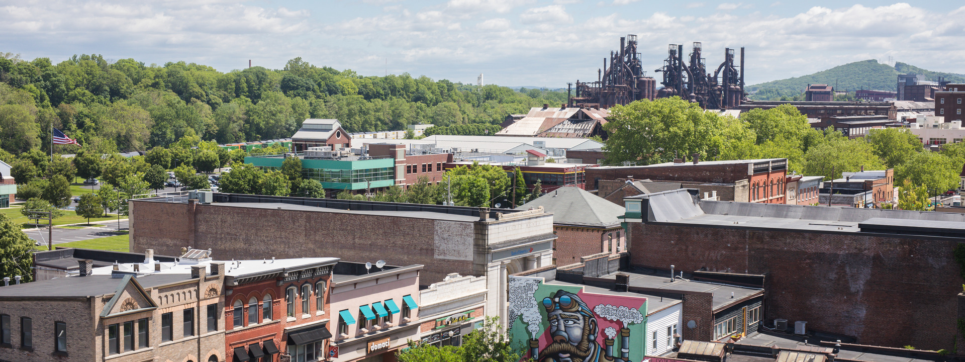 An arial view of South Bethlehem looking east over 2-3 story buildings along third street with larger industrial buildings and a large steel factory in the background. Green trees are throughout the town and on hills in the background, and the sky is blue with light clouds.