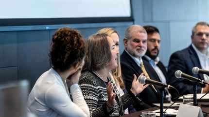 Six panelists seated at a long table with notes and microphones in front of them.