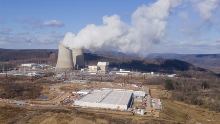 A wide landscape shot depicting a large warehouse-like building with cooling towers of a power plant emitting white clouds of steam behind. A low mountain range is in the distance.