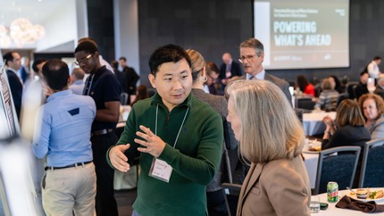 Two people engaged in conversation. They are in a crowded room with many other conversations. A projector screen in the background reads "Powering What's Ahead."