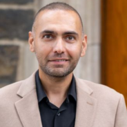 headshot of professor Javad Khazaei, a man with close cut dark hair wearing a light brown jacket over a black shirt post in front of a stone building.